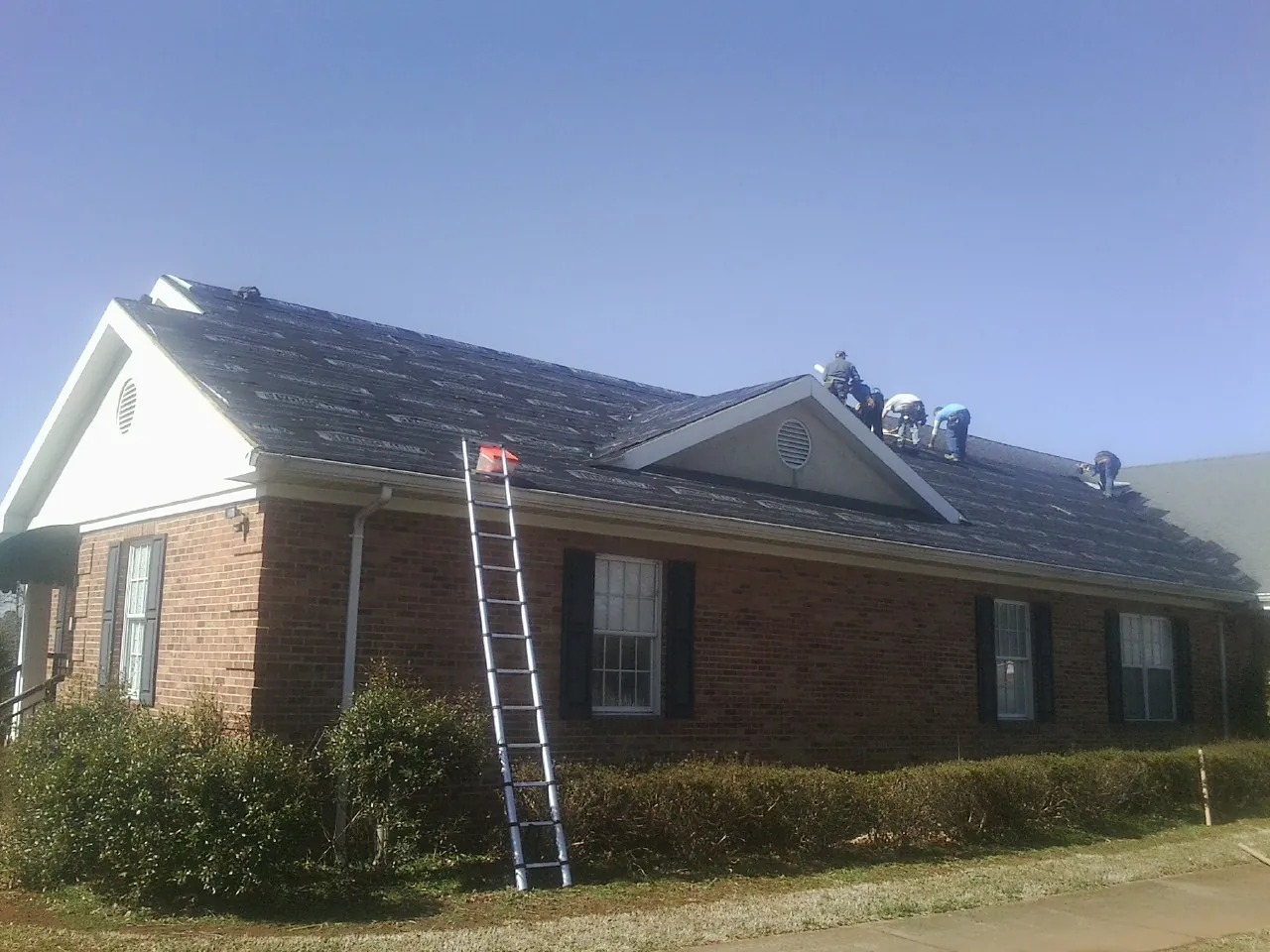 Crew tearing shingles off a brick ranch roof with a ladder set — Emergency Roof Repair work by our crew in Roberts
