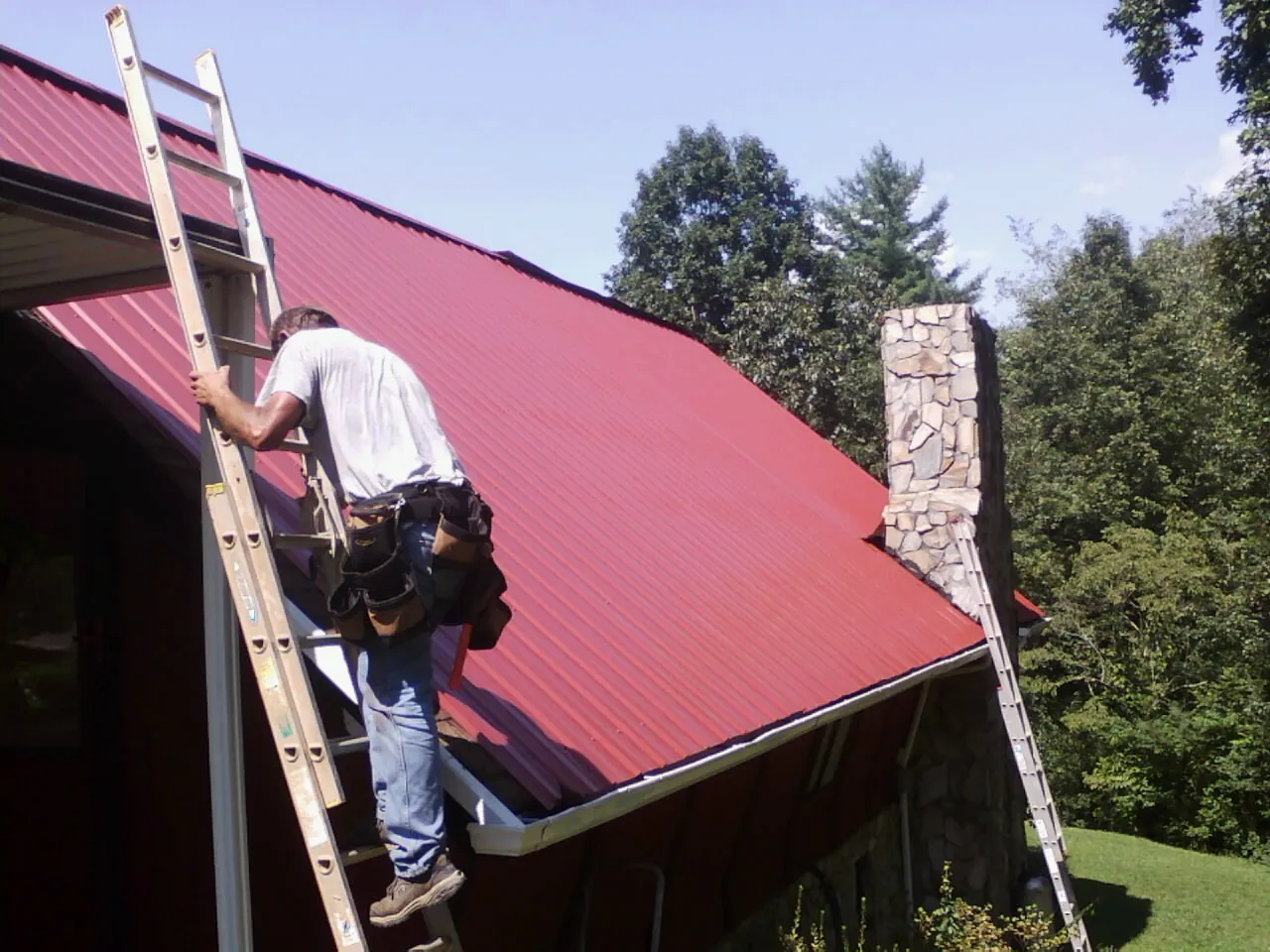 Roofer on a ladder beside a red metal roof and stone chimney — Soffit & Fascia Repair by Roofing Brothers in Roberts